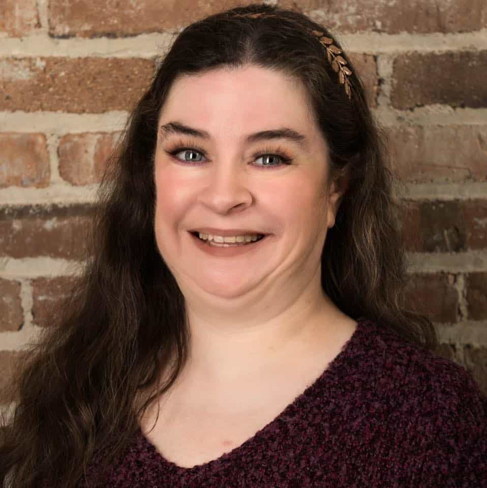 Smiling woman with dark brown hair and blue eyes, brick wall background.